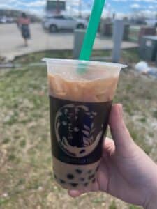A hand holding a Bobablastic iced milk tea with black boba pearls and a green straw outdoors.