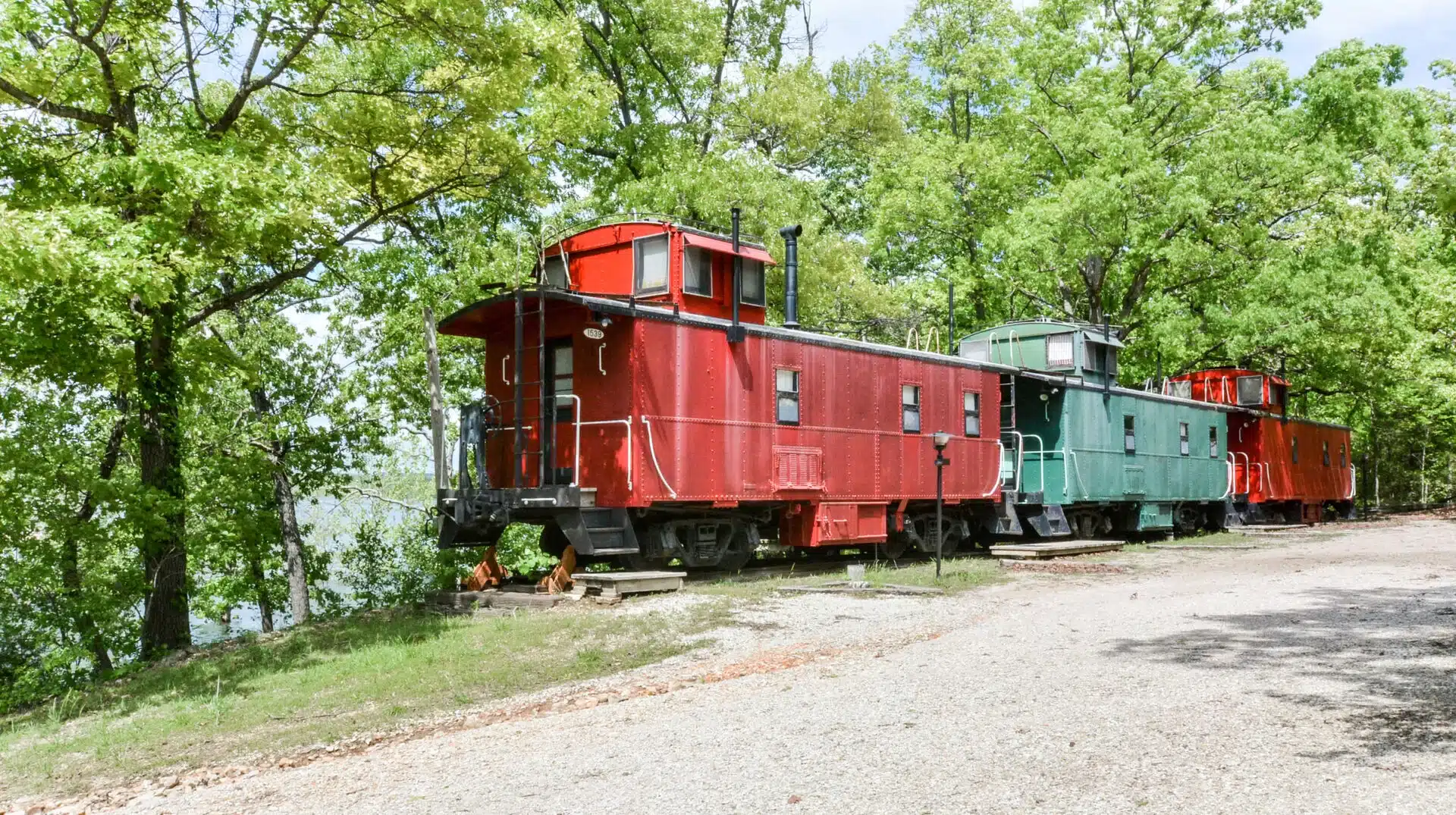 Red and green vintage railroad cabooses lined up under shade trees beside a gravel drive at Caboose Junction Resort in Lampe, MO